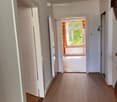 Corridor / Hallway. A clean, well-lit hallway with wooden flooring, white walls, and multiple doors leading to different rooms. Natural light streams in from a window at the end of the hallway.