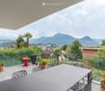 Balcony / Terrace. A modern balcony with a glass railing, offering a scenic mountain view. It features a large dining table with chairs, several potted plants, and a small red side table.
