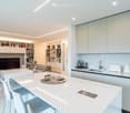 Furnished Room. Modern, open-concept living and dining area with a sleek white kitchen. Features include a large island with bar stools, a built-in bookshelf, a flat-screen TV, and contemporary furniture.
