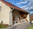 House. Two-story house with beige stone walls, red-tiled roof, and wooden balcony. Features include a small outdoor seating area with a table and chairs, a staircase leading to the balcony, and a paved cobblestone driveway.