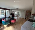 Furnished Room. Modern, well-lit living and dining area with wooden flooring, a glass dining table with colorful chairs, a sofa, and a kitchen counter with appliances.