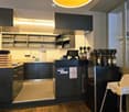 Furnished Room. Modern bar area with dark cabinets, wine bottles, wine glasses, a refrigerator, a table with a screen, and a baby chair.