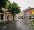 Building. A wet urban street lined with colorful buildings, including a cafe with striped awnings and closed umbrellas, with several parked cars and pedestrians.