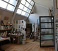 Furnished Room. An artist's studio with wooden floors, large skylights, and various artworks and books displayed on shelves and walls. A desk with art supplies, a chair, and a cabinet are visible.