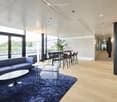 Furnished Room. Modern, well-lit lounge area with a blue sofa, white coffee table, grey armchair, round dining table with stools, and a potted plant near the window.