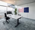 Modern office space with a large white table, ergonomic chairs, computer setup, and a potted plant near large windows offering a city view.