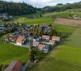 Building. Aerial view of a residential area with multiple houses, surrounded by green fields and forested hills.