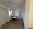 Furnished Room. A small, unfurnished room with wooden flooring, white walls, and a single window. A small table and chairs are present, along with a radiator and a closed door.