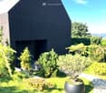 Modern black house with a sloped roof, surrounded by a well-maintained garden with various potted plants and shrubs. The garden features a gravel pathway leading to the house entrance.