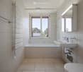 Modern bathroom with a bathtub, sink, toilet, mirror, and showerhead. The room features large windows with a view of the outdoors.