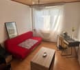 A cozy living room with a red sofa, wooden coffee table, desk with chair, and a chandelier hanging from the ceiling. The room features a large window with white curtains and a framed picture on the wall.