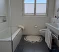 Modern bathroom with a bathtub, toilet, sink, and mirror. Features a round beige bathmat and a dark grey tiled floor.