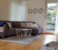 Modern living room with a grey sofa, multiple throw pillows, a small coffee table, a floor lamp, and three wall-mounted clocks. The room features large windows with white curtains and a wooden floor.