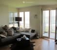 Modern living room with large windows and a sliding glass door leading to a balcony. Features a grey sectional sofa with multiple cushions, a coffee table with decorative items, a floor lamp, and a candle holder near the door.