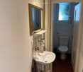 A small bathroom with a sink, mirror, and a toilet visible through an open doorway. The sink has a faucet and a toothbrush holder, and a towel hangs on the wall. A water tank and a green watering can are also present.