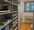 A small, organized room with a large metal shelving unit filled with books and office supplies, a printer on top, a radiator, and a window with blinds.