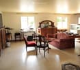 Bright living room with beige walls, tiled floor, large windows, and natural light. Features a red patterned sofa, wooden chairs, a small round table, a wooden cabinet, a mirror, and a lamp.