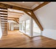 Bright, open loft space with wooden floors, exposed wooden beams, and a staircase leading to an upper level. Features large glass doors and white walls.