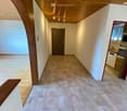 Empty hallway with tiled floor, wooden ceiling, and a closed door at the end. Natural light from the left side and a glimpse of a kitchen area on the right.