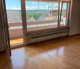 Empty room with wooden flooring, large windows, and a balcony area visible through the open sliding doors.
