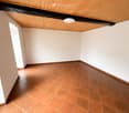 Empty room with white walls, terracotta tiled floor, and a wooden slanted ceiling with exposed beams. A single round light fixture is mounted on the ceiling.