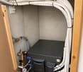 A water pump unit with multiple white pipes, valves, and a pressure gauge installed in a wooden cabinet.