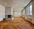 Empty room with wooden herringbone flooring, white walls, large windows, and a fireplace with a built-in bench.