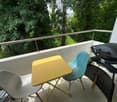 Balcony with a yellow table, two chairs, and a grill, overlooking a green area.