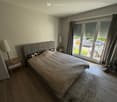 Well-lit bedroom with wooden flooring, large bed with grey headboard and beige bedding, nightstands with lamps, and large windows with curtains leading to a balcony.