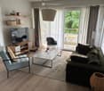 Modern living room with a grey sofa, glass coffee table, light blue armchair, TV, and a balcony with outdoor furniture visible through large windows.