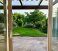 View through a large sliding glass door onto a well-maintained garden with a stone patio, green lawn, and various shrubs and trees.