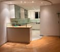 Modern kitchen with white cabinets, glass-fronted upper cabinets, a central island with a glass countertop, and a sleek, minimalist design. The floor is made of light wooden planks, and the space is well-lit with recessed ceiling lights.