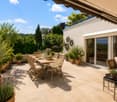 A spacious outdoor patio with a dining table and chairs, a sofa, and several potted plants, surrounded by greenery and a clear blue sky.