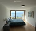Modern bedroom with wooden flooring, large window offering a sea view, bed with blue bedding, nightstand with lamp, and a framed picture on the wall.
