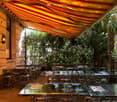 Outdoor seating area with tables and chairs, covered by a large awning, surrounded by greenery and trees.