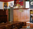 Dining area with wooden tables and chairs, wooden wall paneling, hanging lamps, a broom, a cabinet with shelves, a speaker, a coffee maker, and a bell.