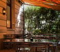 Outdoor seating area with metal tables and chairs, covered by a large awning, surrounded by greenery and a building wall.