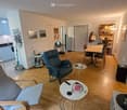 A modern, well-lit living and dining area with wooden flooring, a grey sofa, a blue armchair, a small round table, a dining table with chairs, kitchen cabinets, a refrigerator, a laptop on a small table, a bookshelf, a floor lamp, and a mirror on the wall