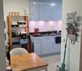 Modern kitchen with light grey cabinets, a wooden dining table with a chair, a coffee maker on the counter, and a shelf with various items.