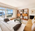 Bright living room with large windows offering a scenic view, featuring a white fur throw on a sofa, a glass coffee table, a wooden bookshelf, and a fireplace.