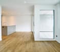 Open-plan unfurnished room with light wooden flooring, white walls, and a small kitchen area visible in the background. The space features large windows and a glass door leading to another room.