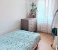Cozy bedroom with a bed covered in a green and white patterned duvet, a wooden nightstand with a lamp, a potted plant, and a wardrobe with hanging clothes.