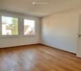 Empty room with light wooden flooring, white walls, and two windows offering a view of neighboring houses and greenery.