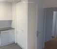 A narrow hallway with white walls, wooden flooring, and a small kitchen area visible on the left side with white cabinets and a countertop.