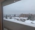 Snow-covered balcony overlooking a residential area with houses, trees, and distant mountains under a cloudy sky.