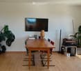 Furnished Room. Bright room with wooden floor, large window, TV screen mounted on wall, wooden dining table with chairs, decorative lamp, and potted plants.