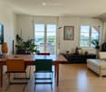 Living Room. Bright living room with wooden flooring, large windows, a cream-colored sofa with pink cushions, a wooden dining table with chairs, a floor lamp, a TV screen, and a fireplace.