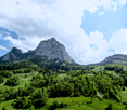 A panoramic view of a lush green valley surrounded by towering mountains under a partly cloudy sky. The landscape features dense forests, rolling hills, and scattered small houses or cabins nestled among the trees.