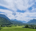 A panoramic view of a lush green valley surrounded by snow-capped mountains, with scattered houses and trees, under a partly cloudy sky.