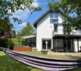 Modern two-story house with white exterior, large windows, balcony, patio area with dining table and chairs, barbecue, fire pit, and hammock in the front yard.